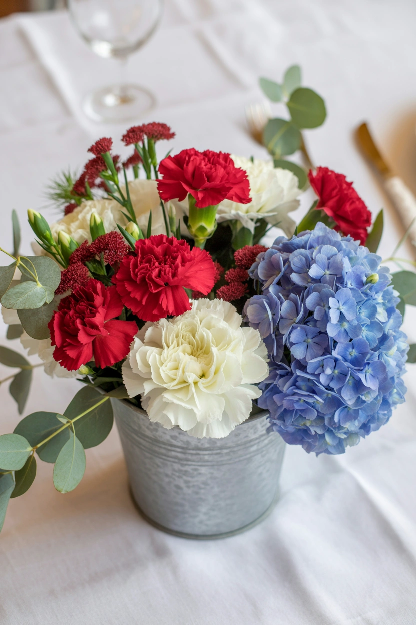 Patriotic 4th of July floral centerpiece with red carnations, white mums, and blue hydrangeas in a galvanized bucket